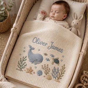 Puede incluir: Una manta de punto para bebé en color crema con bordados de motivos marinos que incluyen una ballena azul, una tortuga marina, estrellas de mar, conchas y algas. El nombre Oliver James está bordado en azul sobre el diseño.