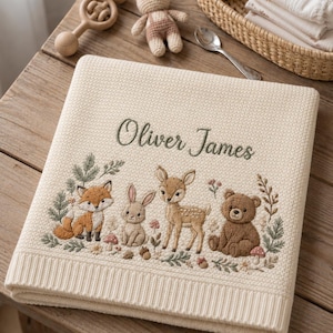 May include: A cream-coloured knit baby blanket featuring an embroidered forest scene with a fox, rabbit, fawn, and bear. The name Oliver James is stitched in dark green script above the animals, surrounded by small floral and mushroom details.