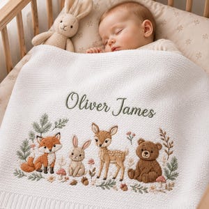 May include: A white knitted baby blanket featuring the embroidered name Oliver James in green script. The blanket displays a detailed woodland scene with a fox, a rabbit, a deer, and a bear surrounded by small flowers, mushrooms, and green foliage.