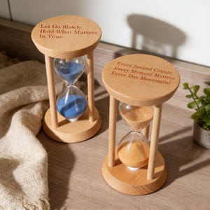 Puede incluir: Dos relojes de arena de madera con citas inspiradoras grabadas. Uno contiene arena azul, el otro arena naranja. El texto del primer reloj de arena dice: "Deja ir lentamente, aferrate a lo que importa en el tiempo". El segundo reloj de arena dice: "Cada segundo cuenta, cada momento importa, cada día es significativo."