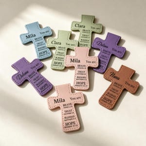 Könnte beinhalten: Eine Sammlung von kreuzförmigen Ornamenten aus Kunstleder in verschiedenen Pastellfarben. Jedes Kreuz trägt einen Namen und inspirierende Worte wie "Brave", "Trust" und "Hope". Die Kreuze sind in Blau-, Lila- und Rosatönen gehalten.