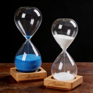 May include: Two glass hourglasses on wooden bases. One contains blue sand, the other white sand. The base of the blue sand hourglass is engraved with "Anastasia". The white sand hourglass base is engraved with "Sublime".