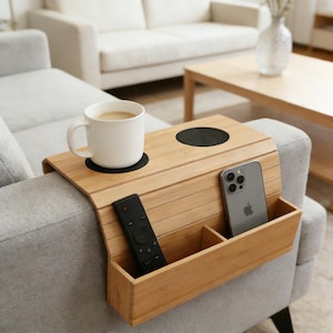 May include: A bamboo sofa arm tray with a built-in cup holder, a small storage compartment, and a larger compartment holding a remote control and a smartphone. A white mug with coffee sits on the tray.