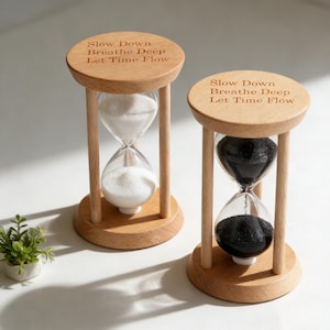 Könnte beinhalten: Zwei Holz-Sanduhren mit der Gravur "Slow Down Breathe Deep Let Time Flow". Eine enthält weißen Sand, die andere schwarzen Sand. Die Sanduhren sind aus hellem Holz gefertigt.