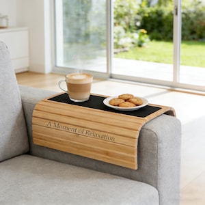 May include: A bamboo sofa arm tray with a black surface, holding a cup of coffee and a plate of cookies. The tray has the text "A Moment of Relaxation" engraved on it. The tray is placed on a gray sofa arm.