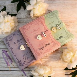 May include: Three pastel-coloured journals with heart-shaped locks and floral designs. The journals are in shades of lavender, pink, and mint green, with the words "Unique & Yours" and "Noble" printed on them. White peonies are scattered around the journals.