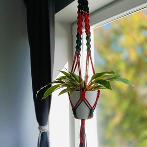 May include: A white ceramic plant pot held by a macrame hanger made of red and dark green cord. The hanger features intricate knotted patterns, a wooden ring at the top for suspension, and a decorative tassel of loose red and green cords at the base.