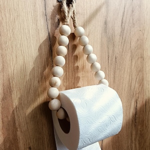 Könnte beinhalten: Ein Toilettenpapierhalter bestehend aus einer Schlaufe aus glatten, hellen Holzperlen, die auf einer Schnur aus natürlicher Jute aufgefädelt sind. Der Halter hängt an einem kleinen, dunklen Metallhaken und trägt eine Rolle weißes Toilettenpapier.