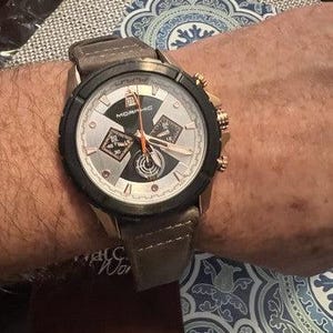 May include: A gold-toned wristwatch with a black bezel and a light-colored face. The watch has a brown leather band and features multiple dials and hands. The watch is on a wrist, with a patterned background visible.