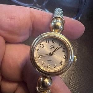 May include: A gold-toned quartz watch with a round face and a silver dial. The watch features black numerals and hands, with the brand name "Cale" printed on the dial. The watch has a braided band.