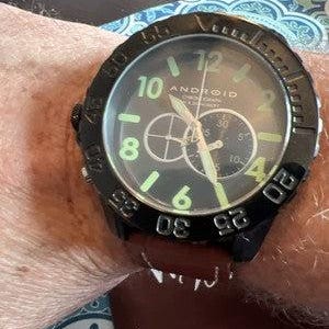 May include: Black Android chronograph watch with a brown leather strap. The watch face is black with white and green detailing, including the numerals and hands. The watch features two sub-dials and the word "ANDROID" is printed on the face.