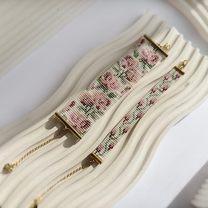 May include: Two beaded bracelets with floral patterns in pink, green, and white. The larger bracelet is 1.5 inches wide, and the smaller one is 0.75 inches wide. Both have gold-colored clasps and chains. The bracelets rest on a white, wavy surface.