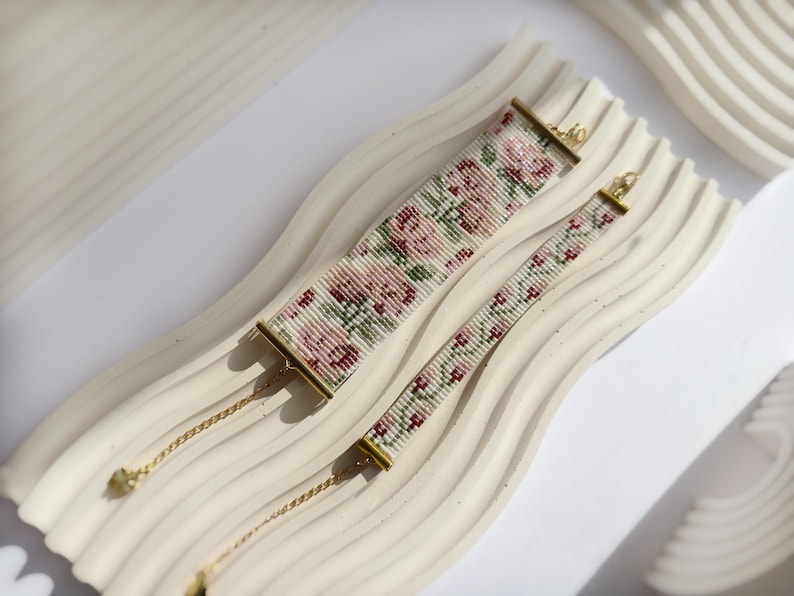 May include: Two beaded bracelets with floral patterns in pink, green, and white. The larger bracelet is 1.5 inches wide, and the smaller one is 0.75 inches wide. Both have gold-colored clasps and chains. The bracelets rest on a white, wavy surface.