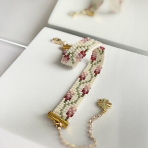 May include: A beaded bracelet with a floral pattern in cream, pink, and green. The bracelet has gold-colored metal clasps and a small chain with a four-leaf clover charm. The bracelet is approximately 6 inches long.