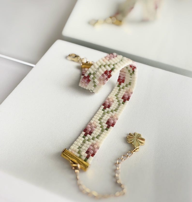 May include: A beaded bracelet with a floral pattern in cream, pink, and green. The bracelet has gold-colored metal clasps and a small chain with a four-leaf clover charm. The bracelet is approximately 6 inches long.