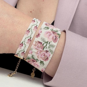 May include: Two beaded bracelets with a floral design. The bracelets feature a white background with pink and green flowers. The bracelets are connected with gold-colored chains and clasps. The bracelets are worn on a wrist.