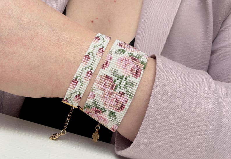 May include: Two beaded bracelets with a floral design. The bracelets feature a white background with pink and green flowers. The bracelets are connected with gold-colored chains and clasps. The bracelets are worn on a wrist.