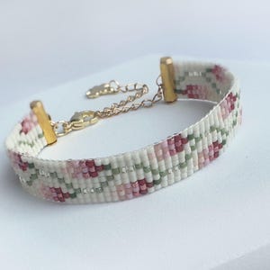 May include: A beaded bracelet with a floral pattern in shades of pink, green, and white. The bracelet has gold-colored metal accents and a chain for adjustable sizing. The bracelet is a delicate piece of jewelry.