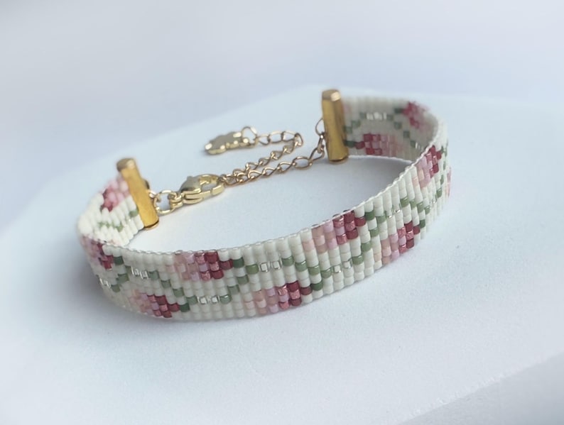 May include: A beaded bracelet with a floral pattern in shades of pink, green, and white. The bracelet has gold-colored metal accents and a chain for adjustable sizing. The bracelet is a delicate piece of jewelry.