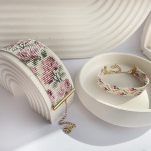 May include: Two beaded bracelets with floral patterns. One is displayed on a white arch-shaped stand, the other in a white bowl. The bracelets feature pink, green, and white beads, with gold-colored accents and chain details.