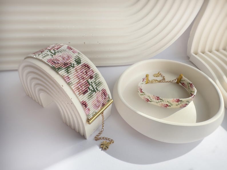 May include: Two beaded bracelets with floral patterns. One is displayed on a white arch-shaped stand, the other in a white bowl. The bracelets feature pink, green, and white beads, with gold-colored accents and chain details.