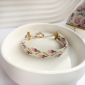 May include: A beaded bracelet with a floral pattern in shades of pink, green, and burgundy on a white background. The bracelet has gold-colored metal accents and a delicate chain. A similar beaded piece is also visible.