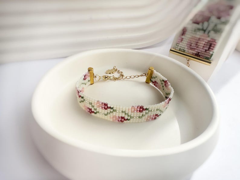 May include: A beaded bracelet with a floral pattern in shades of pink, green, and burgundy on a white background. The bracelet has gold-colored metal accents and a delicate chain. A similar beaded piece is also visible.