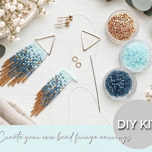 Può includere: Un kit fai-da-te per creare orecchini a frange con perline, composto da due telai metallici triangolari, perni dorati, anellini, un ago, filo trasparente e tre piccoli contenitori circolari riempiti con perline dorate, azzurre e blu scuro.