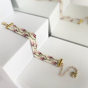 May include: A beaded bracelet with a floral pattern in shades of pink, green, and white. The bracelet has gold-colored metal accents and a delicate chain with a flower charm. The bracelet is displayed on a white surface.
