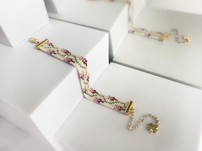 May include: A beaded bracelet with a floral pattern in shades of pink, green, and white. The bracelet has gold-colored metal accents and a delicate chain with a flower charm. The bracelet is displayed on a white surface.
