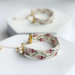May include: Two beaded bracelets with floral patterns. The bracelets feature a cream-colored base with pink, red, and green floral designs. Gold-colored clasps and adjustable chains complete the design. One bracelet is in focus, the other is blurred in the background.