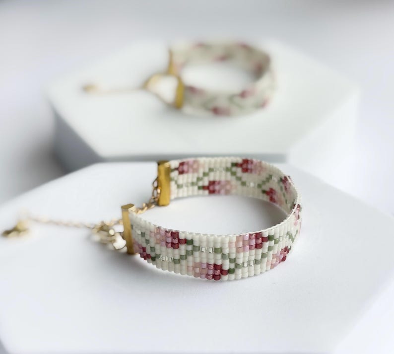 May include: Two beaded bracelets with floral patterns. The bracelets feature a cream-colored base with pink, red, and green floral designs. Gold-colored clasps and adjustable chains complete the design. One bracelet is in focus, the other is blurred in the background.