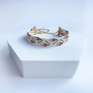May include: A beaded bracelet with a floral pattern in shades of pink, green, and white. The bracelet has gold-colored metal accents and a chain for adjustable sizing. The bracelet is displayed on a white geometric stand.