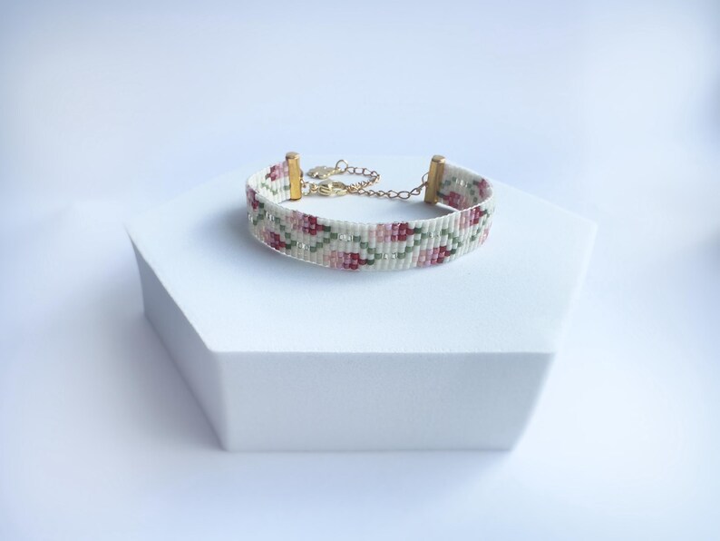 May include: A beaded bracelet with a floral pattern in shades of pink, green, and white. The bracelet has gold-colored metal accents and a chain for adjustable sizing. The bracelet is displayed on a white geometric stand.