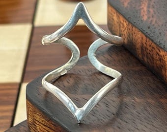 May include: A silver-colored ring with a geometric design, featuring a series of sharp angles and curves. The ring is displayed on a dark wooden surface, with a chessboard and chess pieces in the background.