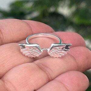 May include: A silver ring featuring a pair of detailed wings. The wings are positioned on either side of the ring's band, creating a unique and symbolic design. The ring is shown in a close-up shot.