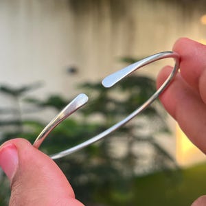 May include: A silver-toned open cuff bracelet with a smooth, polished finish. The bracelet features a slender, rounded band that tapers slightly toward the two flat, rounded ends, creating a simple and continuous circular shape.