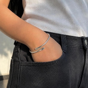 Peut inclure: Un bracelet manchette argenté avec un design ouvert. Le bracelet épouse le poignet, les extrémités se chevauchant. Le bijou est porté sur le bras d'une personne, avec un haut blanc et un jean noir en arrière-plan.