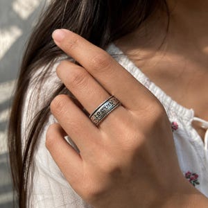 May include: A silver-toned metal ring featuring an embossed band with intricate script characters and a rope-style border along the top and bottom edges. The band has a dark, oxidized finish within the recessed areas to highlight the engraved details.