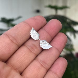 May include: A pair of silver-colored wing-shaped earrings. Each earring features detailed feather designs, with the wings connected by a small bar. The earrings are displayed against a blurred background.