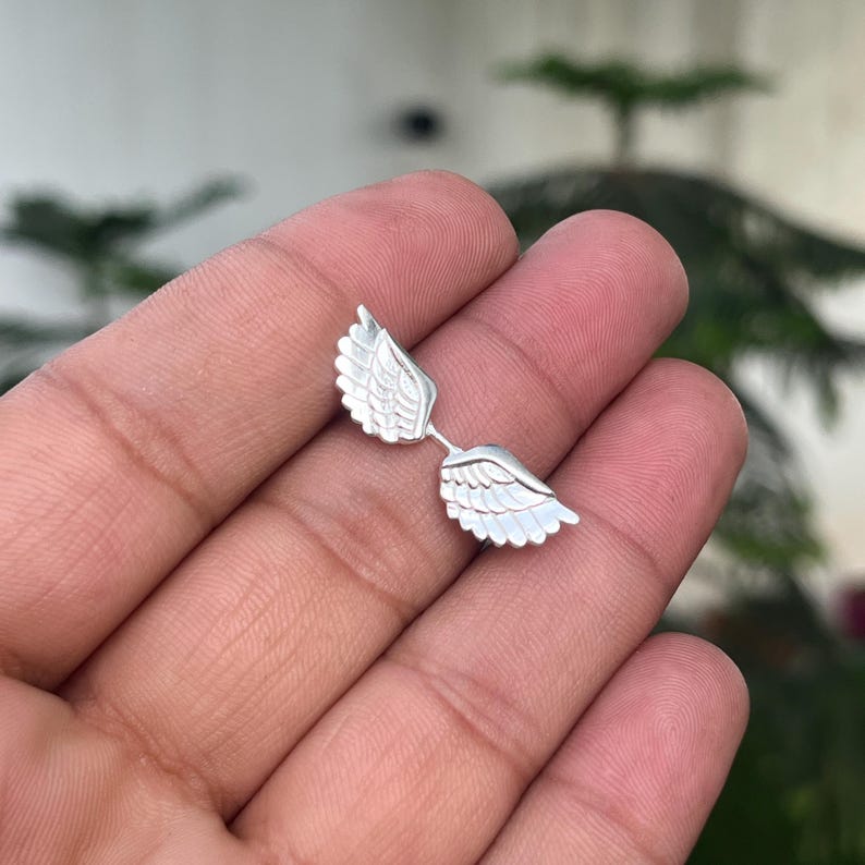 May include: A pair of silver-colored wing-shaped earrings. Each earring features detailed feather designs, with the wings connected by a small bar. The earrings are displayed against a blurred background.