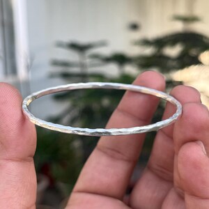 Puede incluir: Un brazalete de plata con una textura martillada. El brazalete es redondo y parece estar hecho de metal. El brazalete se sostiene en la mano de una persona contra un fondo borroso de vegetación.