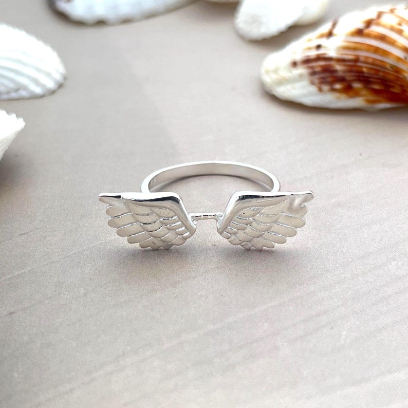 May include: A silver ring featuring a pair of detailed angel wings. The ring is set against a neutral background with seashells, highlighting the ring's design. The wings are symmetrically placed, creating a delicate and symbolic piece of jewelry.