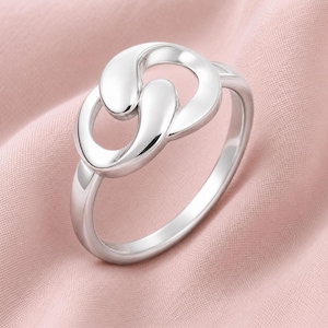 Può includere: Un anello in argento lucido caratterizzato da un design centrale composto da due anelli intrecciati a forma di goccia liscia. La fascia è solida e arrotondata, mantenendo una finitura metallica uniforme su tutta la struttura del gioiello.