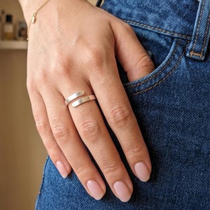 May include: A silver-colored wrap ring with a minimalist design. The ring features two parallel bands that overlap slightly, creating an open, adjustable style. The ring is worn on a finger, with a denim background.