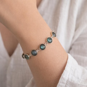 May include: A silver bracelet featuring round, iridescent, blue-green gemstones. The bracelet is composed of circular bezel-set stones linked together. The jewelry is displayed on a person's arm, against a white background.