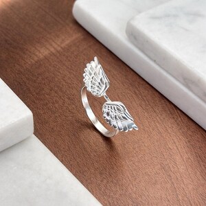 May include: A silver ring featuring an open design with two detailed wing motifs. The ring is set against a brown and white background, showcasing the intricate feather detailing and the ring's polished finish. The ring is a piece of jewelry.