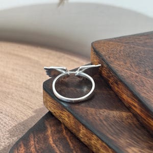 May include: A silver ring with a wing design. The ring features two stylized wings extending outwards from the band. The ring is displayed on a dark wooden surface.