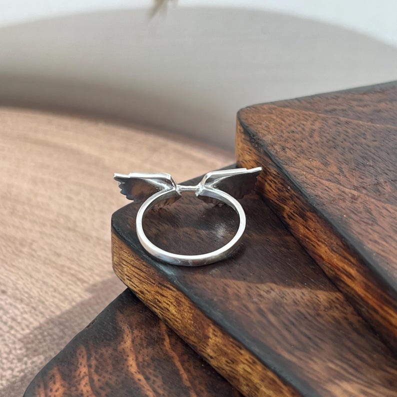 May include: A silver ring with a wing design. The ring features two stylized wings extending outwards from the band. The ring is displayed on a dark wooden surface.