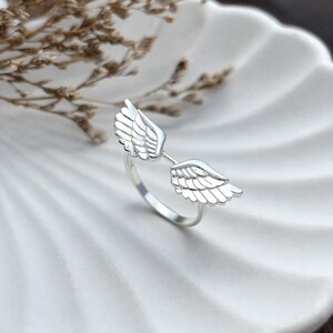 May include: A silver ring featuring a pair of detailed angel wings. The wings are positioned on either side of the ring's band, creating an open design. The ring is displayed on a white, textured surface.